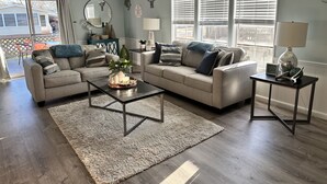 A living room with two sofas, a coffee table, and a side table with a lamp. There are decorative wall pieces and a view of the outside. - Coastal cottage minutes from the beach.  Large front porch for morning sun!! (Myrtle Beach)