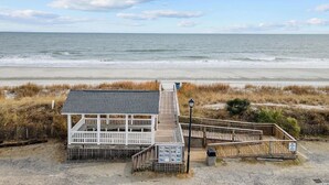A boardwalk leading to a white building with a dark roof, situated on a beach with dunes and the ocean in the background.