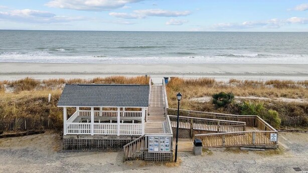 A boardwalk leading to a white building with a dark roof, situated on a beach with dunes and the ocean in the background. - Coastal cottage minutes from the beach.  Large front porch for morning sun!! (Myrtle Beach)