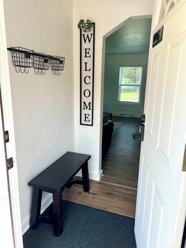 A hallway with a 'WELCOME' sign, a bench, a small shelf with decorative items, and a door leading to another room.