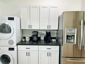 Una cocina con armarios blancos, cafetera, refrigerador y lavadora.