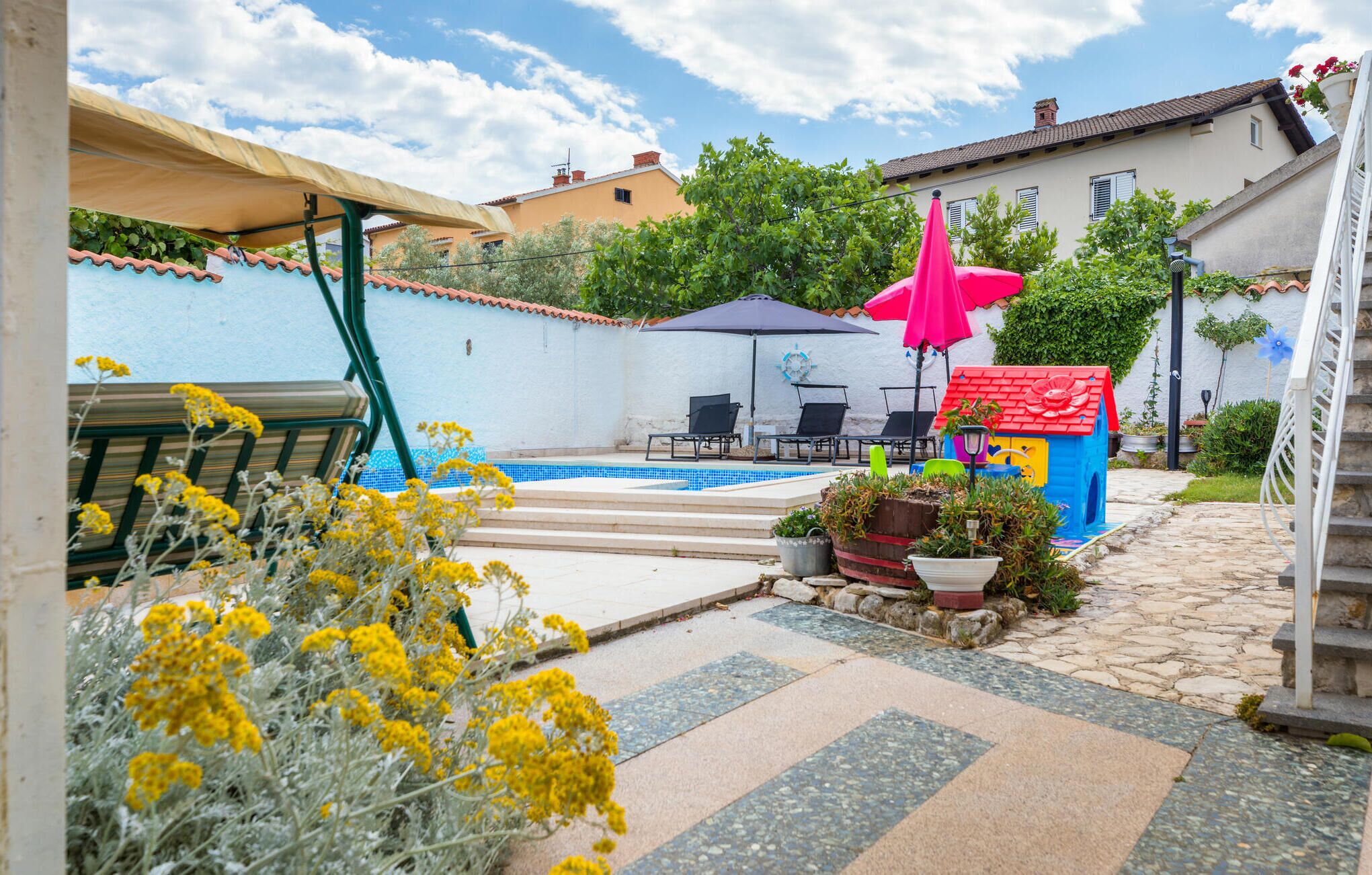 Ein Poolbereich mit einem blau gefliesten Pool, zwei Liegestühlen, einem roten Sonnenschirm und einem kleinen, blauen Spielhäuschen.