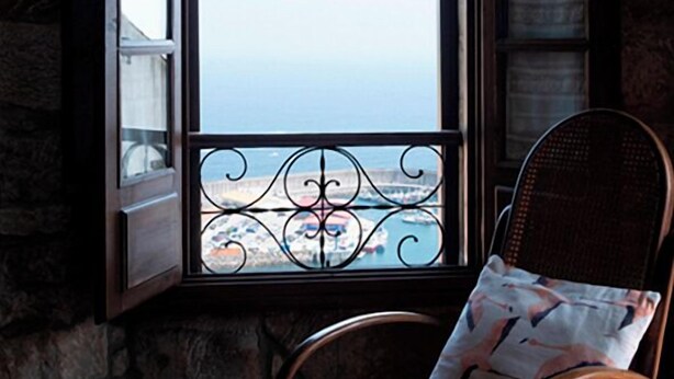 A room with a stone wall, a window with a view of the sea, and a wicker chair with a cushion.