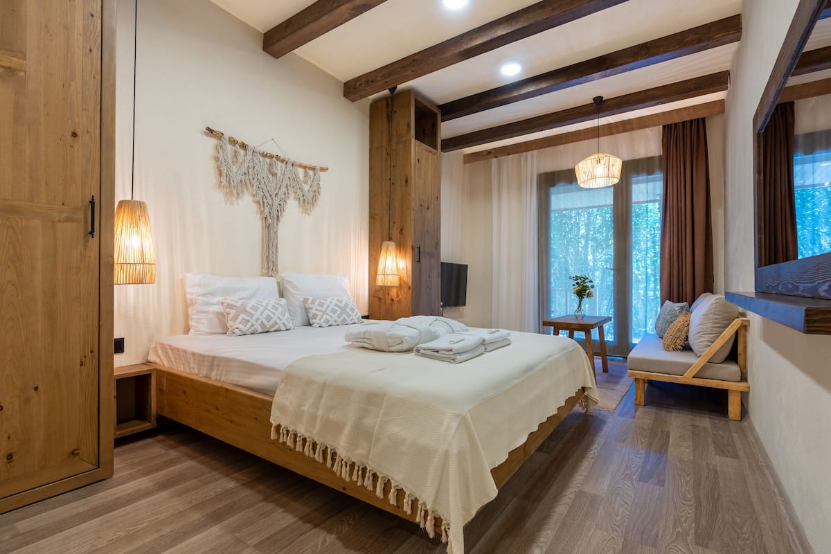 Un dormitorio con una cama de madera, lámparas de noche, un banco de madera y un amplio ventanal con cortinas.