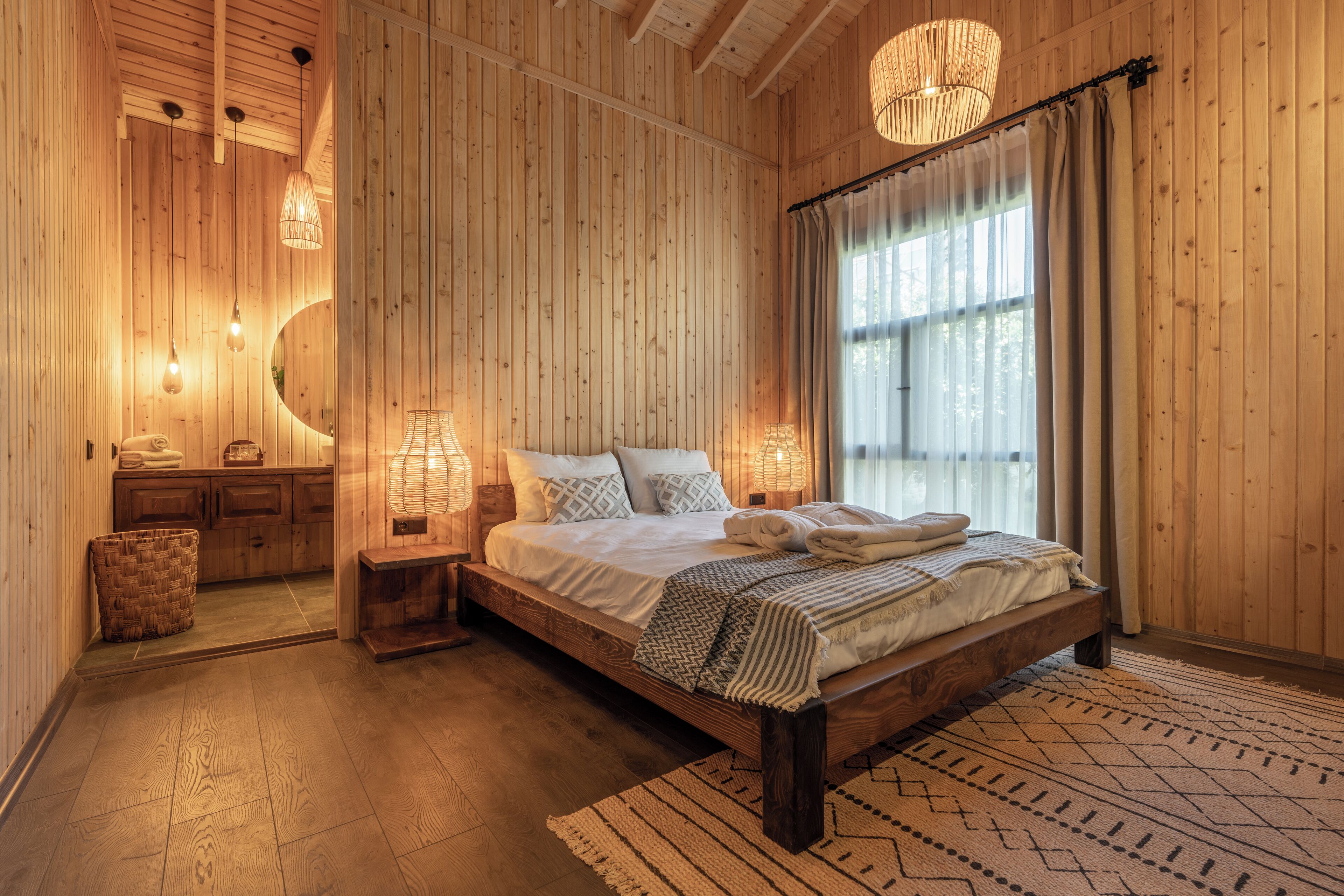 Quarto de madeira com cama, mesas de cabeceira, espelho e uma janela grande com cortinas.