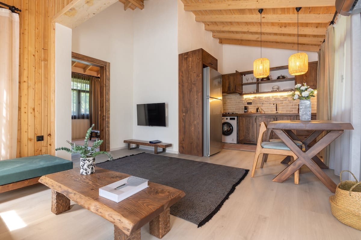 Una sala moderna con un banco de madera, una mesa de centro y un televisor. Se ve una cocina al fondo con armarios de madera y una mesa de comedor.