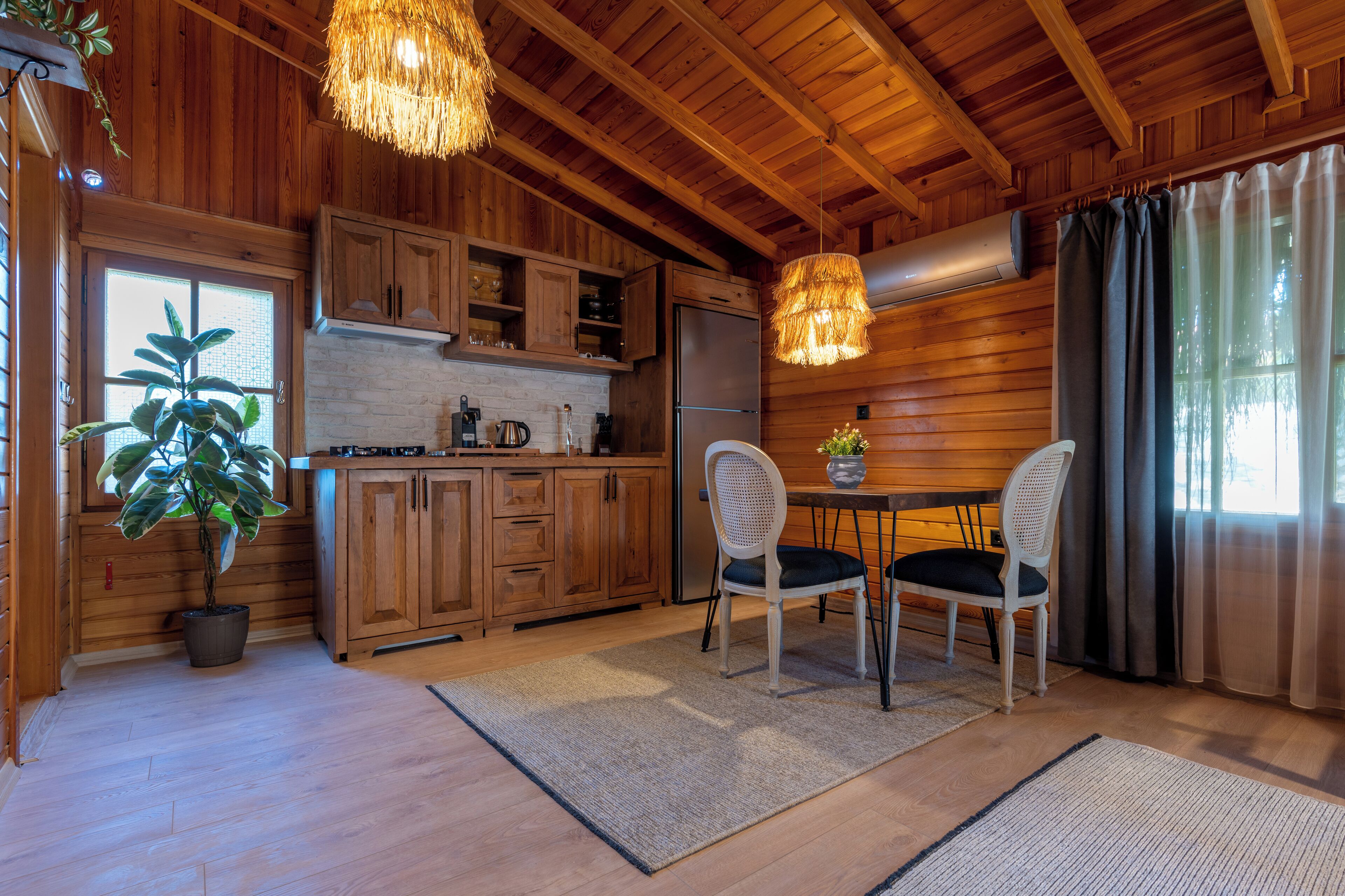 A wooden cabin interior with a kitchen, dining area, and a large window with curtains.