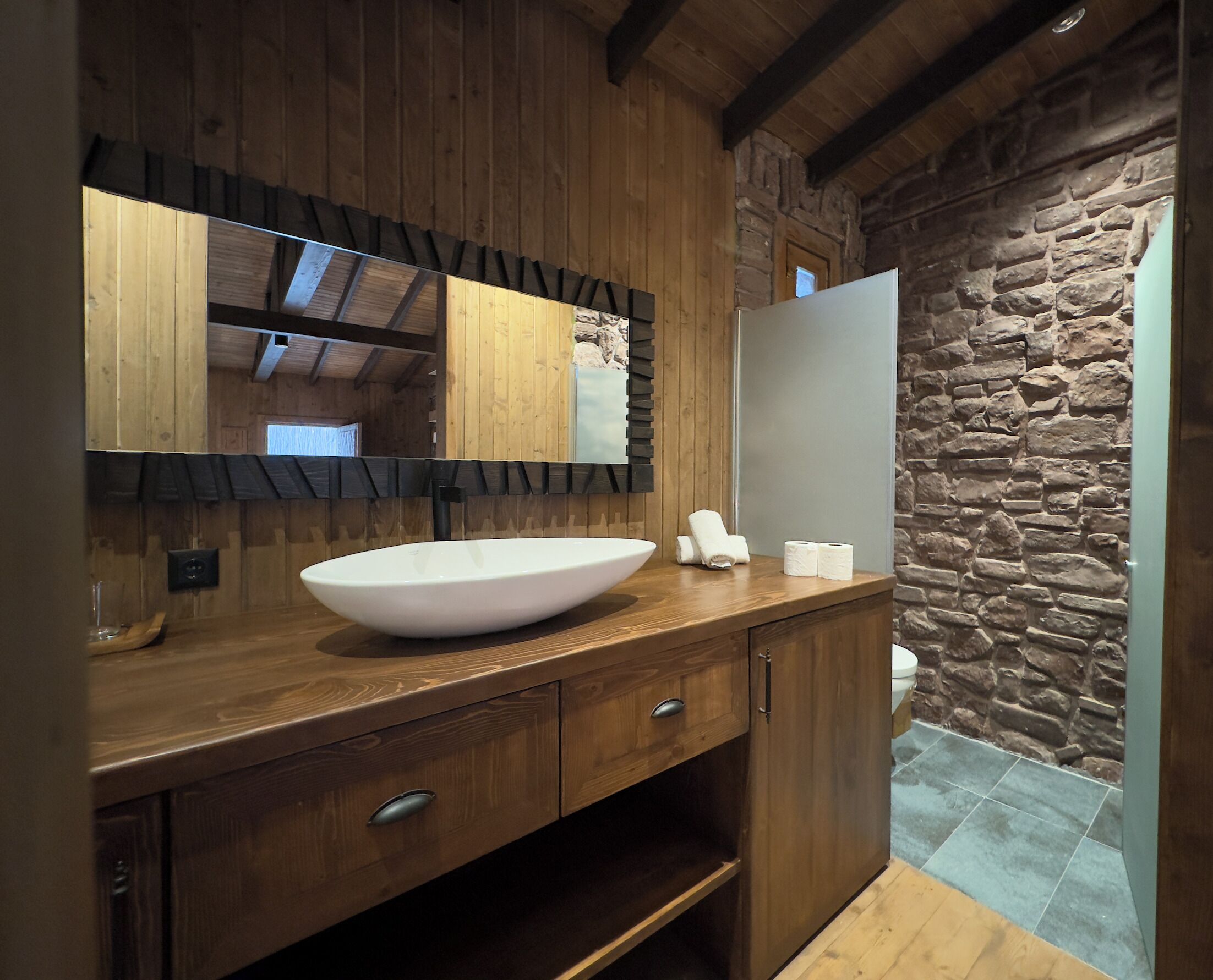 A bathroom with a wooden vanity, a large mirror, a stone wall, and a white sink.