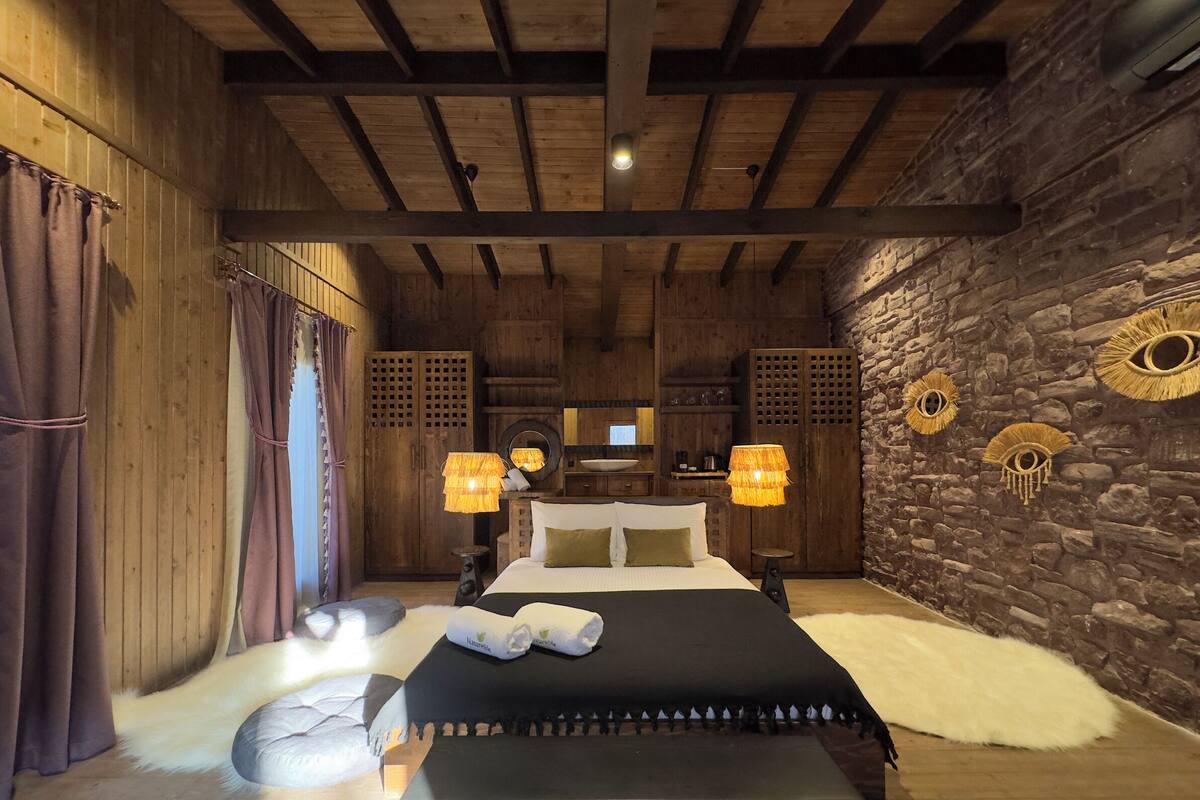 Un dormitorio rústico con techo de madera, paredes de piedra, cama con ropa blanca y dos lámparas con pantallas tejidas.