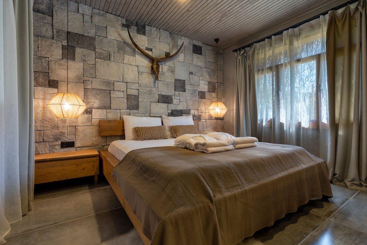 Un dormitorio con pared de piedra, una cama grande con manta marrón, dos lámparas de noche y un banco de madera.
