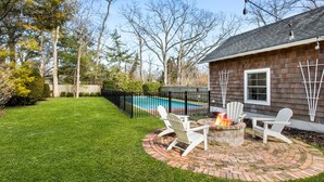 A backyard with a brick fire pit, white Adirondack chairs, and a swimming pool.