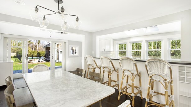 A spacious dining area with a marble table and wicker chairs, large windows with a view of the outdoors, and a modern chandelier.