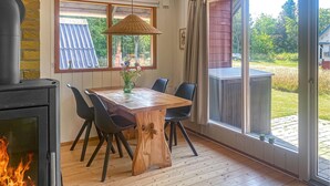 Een houten eettafel met stoelen, een open haard en een groot raam met uitzicht op de natuur.