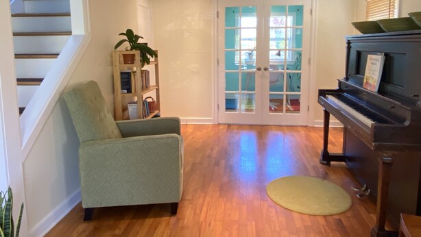 A living room with a piano, a green armchair, a bookshelf, and a door with glass panels. - Charming, light-filled SE Eugene 2-story house  (Eugene)