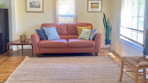 A living room with a brown leather sofa, two side tables, a large woven rug, and two windows with blinds. - Charming, light-filled SE Eugene 2-story house  (Eugene)