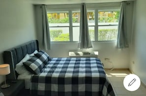 A bedroom with a bed, a nightstand, a window with curtains, and an air conditioning unit. - Beautiful 2 bedrooms condo steps away from the beach (Loíza)