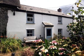 A white building with multiple windows and a door, surrounded by a garden with various plants and flowers.