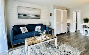 Een woonkamer met een blauwe bank, een salontafel, een schilderij aan de muur en een witte deur met luiken.