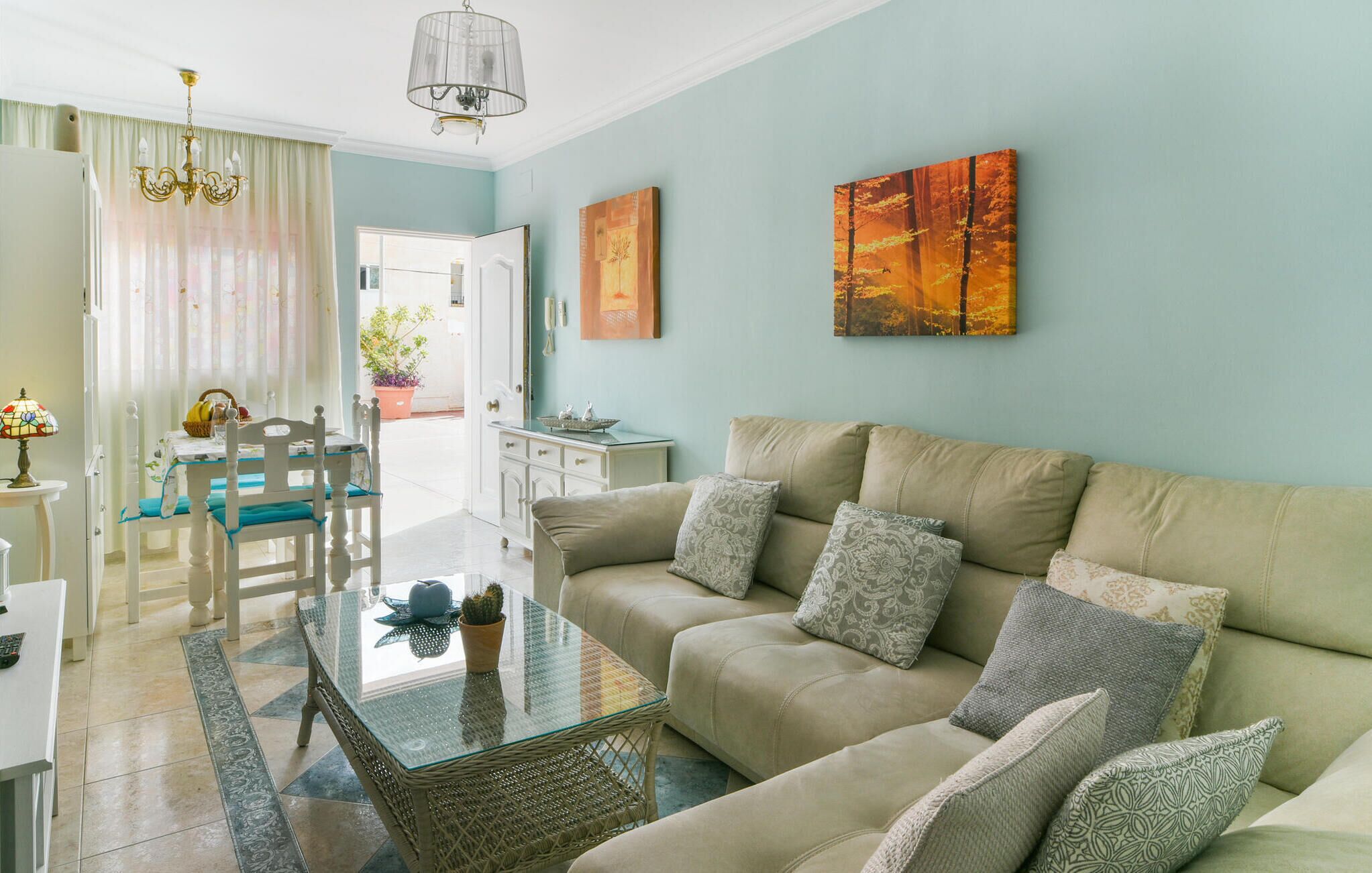 A living room with a beige sofa, a glass coffee table, and a dining area with a white table and chairs. There are two paintings on the wall and a chandelier above the dining table.