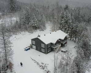 Une grande cabane à deux étages, dont le toit est recouvert de neige, et qui est entourée d’une forêt.