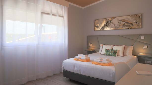 A neatly arranged hotel room with a bed, bedside tables, and a large window with sheer curtains. - Parrots Village (Ribeira Grande)