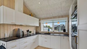 Een moderne keuken met witte kasten, een zwart aanrecht en een rvs afzuigkap. Er is een raam met uitzicht op de buurt, en een hangende oranje pendelarmatuur.