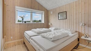 Een slaapkamer met een houten lambrisering, een groot raam met uitzicht op groen, een bed met witte beddengoed en een klein plankje met een beeldje.