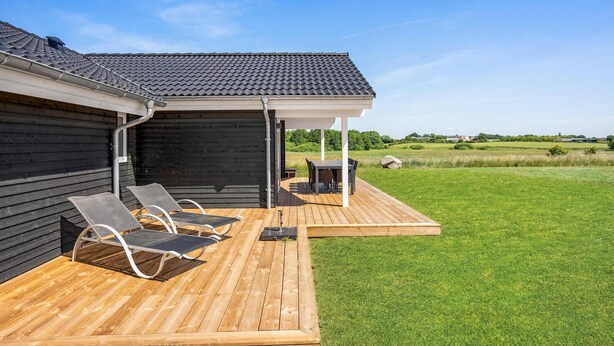 Een houten terras met twee loungesteunen en een tafel, een overdekte patio en uitzicht op een groen veld en verre gebouwen.