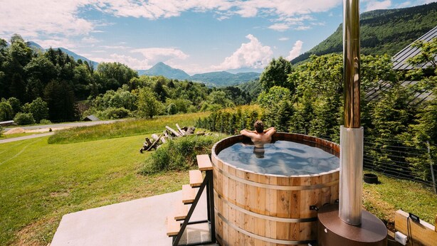 A wooden hot tub with a metal chimney, surrounded by a lush green landscape and mountains in the background. - CHALET DE LA FOUGERE 15 PERS 180M2 BAUGES (Thoiry)