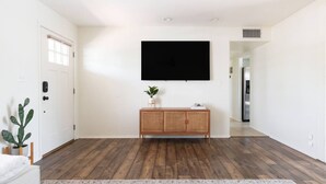Een ruime woonkamer met houten vloer, een grote flatscreen-televisie aan de muur, een rieten console tafel met een plant, en een kamerkleed met dessin.