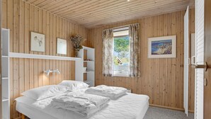 Ein Schlafzimmer mit Holzwandverkleidung, einem Bett mit weißer Bettwäsche, einem Nachttisch, einem Regal mit Dekoration und einem Fenster mit Vorhängen.