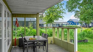 Eine überdachte Terrasse im Freien mit Tisch und Stühlen, umgeben von Grünpflanzen und Blick auf Häuser im Hintergrund.
