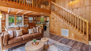 Un interior espacioso de madera con techo alto, una escalera, una sala de estar con sofá y mesa de centro, y una cocina al fondo.