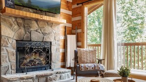 Un salón acogedor con chimenea de piedra, un sillón cómodo, una mesa de centro redonda de madera y vistas al exterior a través de amplias ventanas.
