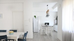 Una cocina moderna con muebles blancos, una zona de comedor con mesa blanca y sillas azules, y una lámpara colgante.