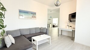 Una sala de estar moderna con un sofá seccional gris, una mesa de centro blanca, un cuadro de un paisaje, un área de comedor con una mesa y sillas, y una cocina con un televisor.