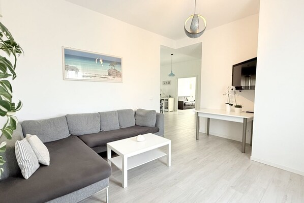 A modern living room with a grey sectional sofa, a white coffee table, a painting of a landscape, a dining area with a table and chairs, and a kitchen area with a TV.