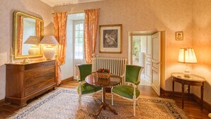 Ein Zimmer mit einem Holzkoffer, zwei grünen Polsterstühlen, einem Glastisch und einem kleinen runden Tisch mit Lampe.