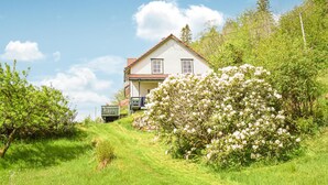 Een huis met twee verdiepingen en een schuin dak, omgeven door weelderig groen en bloeiende struiken.