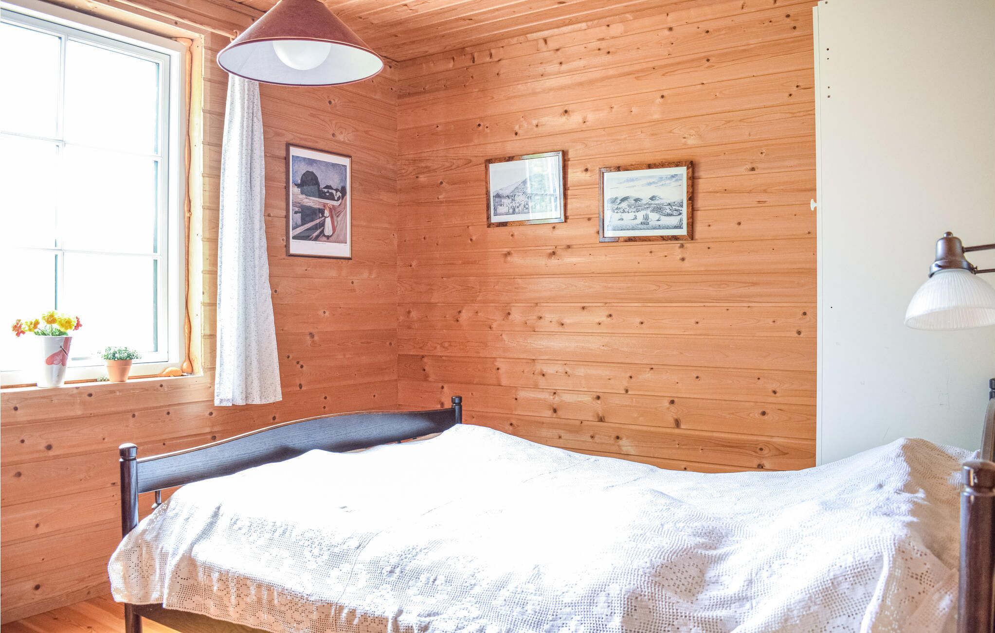 A wooden-paneled room with a bed, a window with curtains, and two framed pictures on the wall.