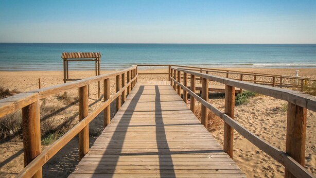 Un malecón de madera que lleva a una playa con un banco y un puesto de salvavidas.