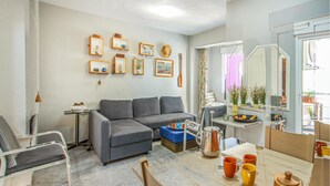 Una sala moderna con un sofá seccional gris, una mesa de comedor con tazas anaranjadas y una zona de cocina con hervidor y frutero.