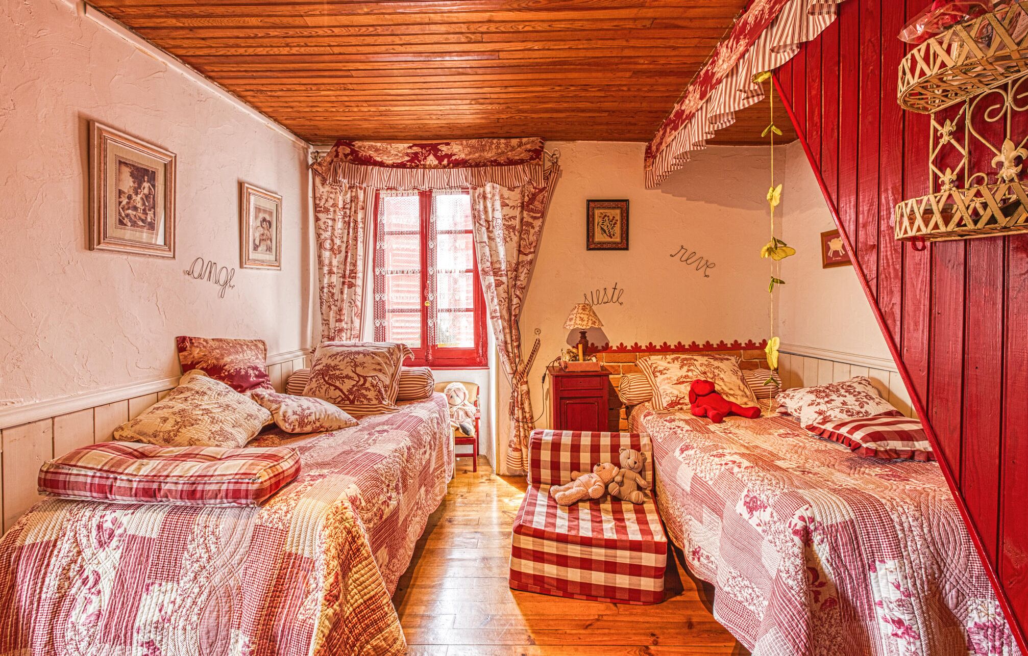 A cozy bedroom with two beds, a window with curtains, and a red wall with decorative elements.