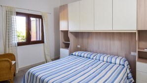 A bedroom with a bed, a chair, a window with curtains, and built-in cabinets. - 1 bedroom beach front home in Budoni (Budoni)