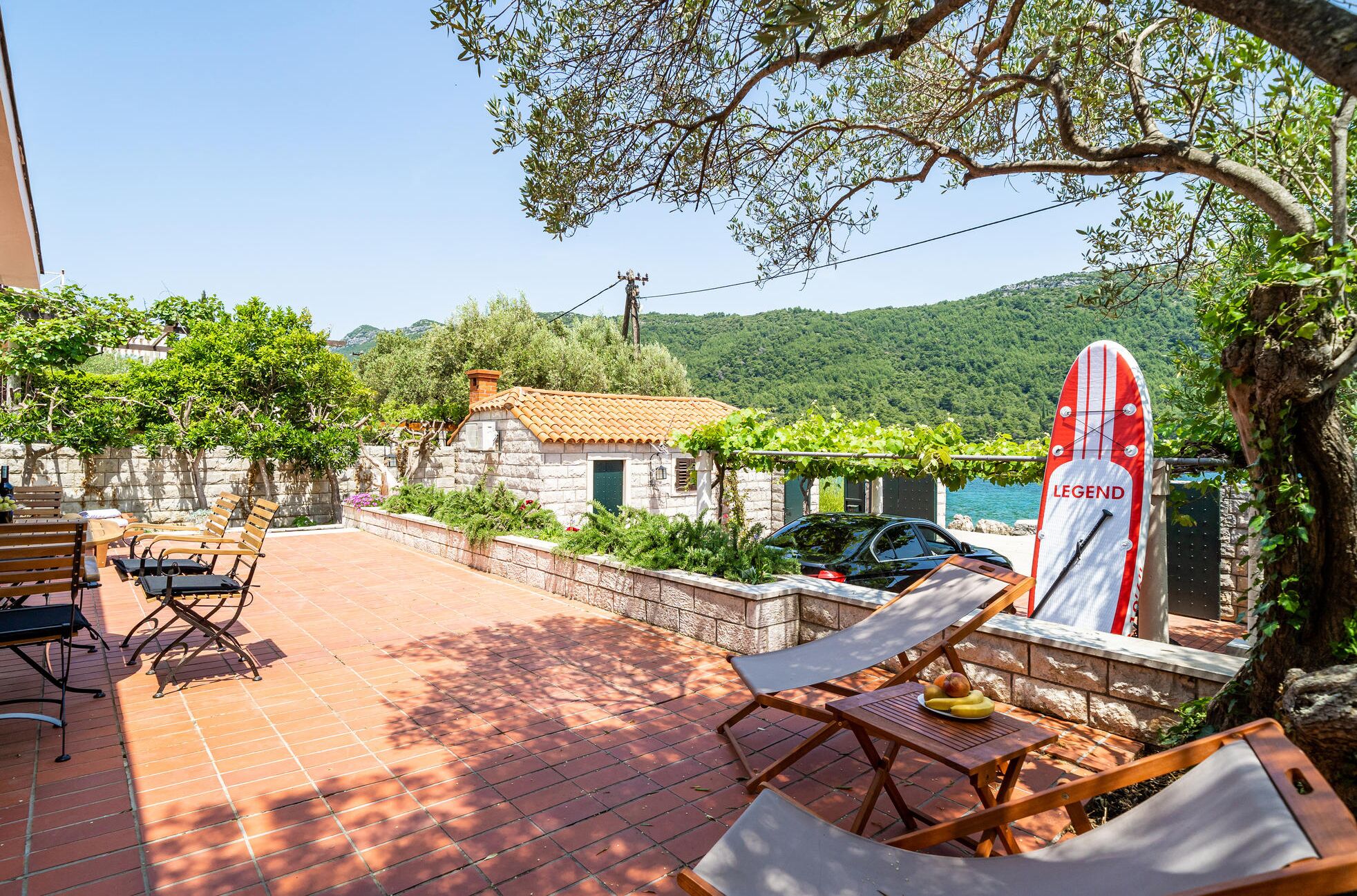Eine Terrasse mit Gartenmöbeln, eine Steinmauer und Blick auf einen See und Berge.