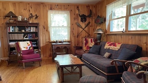 Ein gemütliches Blockhaus-Interieur mit Holzwänden, einem karierten Sofa, einem Schaukelstuhl, einem Holztisch und einem Bücherregal voller Bücher.
