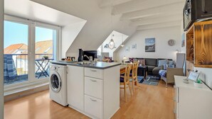 Une cuisine moderne équipée d’un lave-linge intégré, un coin repas avec une table et des chaises, et un salon avec un canapé et un téléviseur.