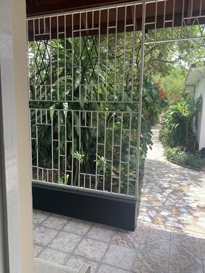 Un espacio al aire libre techado con piso de baldosas y una reja metálica que da a un sendero en el jardín.