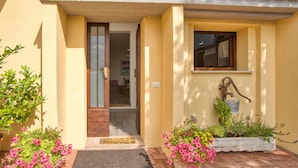A yellow building with a wooden door, a window, and a water pump. There are potted plants and flowers in front.