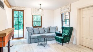 Una habitación con un sofá gris, un sillón verde, una mesita redonda pequeña y un banco de madera. Hay dos ventanas con cortinas, una lámpara de techo y una puerta.
